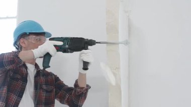 Kırmızı kareli gömlek, mavi başlık ve koruyucu eldiven giyen inşaat işçisi, döner çekiç matkabıyla beyaz duvarı yıkıyor, toz ve portre görüntüsü yaratıyor. Yenileme konsepti.