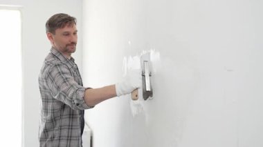 Eldiven ve ekose gömlek giyen orta yaşlı inşaat işçisi, yenileme projesi sırasında pürüzsüz bir yüzey oluşturmak için beyaz duvar alçılamak için son malayı kullanıyor..