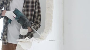 Tanınmayan bej kareli gömlek ve koruyucu eldivenler giyen erkek inşaat işçisi, döner çekiç matkabıyla beyaz duvarı yıkıyor, toz üretiyor, yakın görüş sağlıyor. Yenileme konsepti.