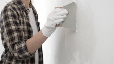 Bilinmeyen, koruyucu eldiven ve kareli gömlek giyen kadın inşaat işçisi, yenileme projesi sırasında beyaz duvara alçı yayıyor, macun bıçağıyla pürüzsüz yüzey.