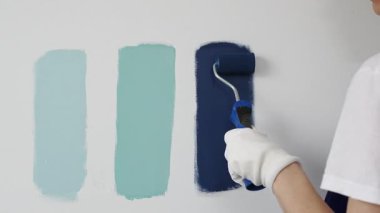 Eldiven giyen ressam beyaz bir duvara boya silindiri uyguluyor, mavi rengin çeşitli tonlarını test ederek iç tasarım için güzel bir eğim etkisi yaratıyor.