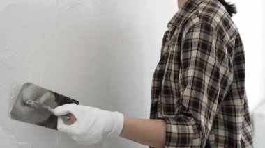 Koruyucu eldiven ve kareli tişört giyen kimliği belirsiz kadın inşaat işçisi, beyaz duvarda ıslak alçıyı düzleştiriyor ve iç dekorasyon sırasında profesyonel malayla malzeme yayıyor.