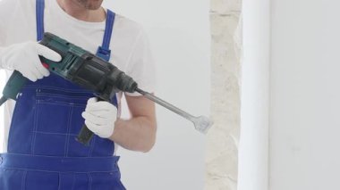 Mavi tulumlu ve koruyucu beyaz eldivenli tanımlanamayan erkek inşaat işçisi, beyaz duvarı döner çekiç matkapıyla yıkıyor, toz üretiyor ve yatay görüşü kapatıyor. Yenileme