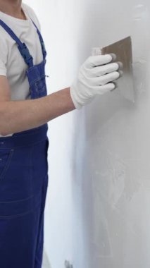 Meçhul inşaat işçisi beyaz eldiven ve mavi tulum giyen kadın, macun bıçağı kullanarak beyaz bir duvara alçı sürüyor. Ev yenileme projesi sırasında pürüzsüz bir yüzey oluşturuyor.