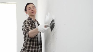 Koruyucu eldiven ve kareli gömlek giyen kadın, iç dekorasyon çalışmalarını boyamadan önce hasarlı bölgeleri yumuşatmak için beyaz duvar yüzeyine macun sürüyor..
