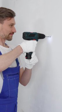 Mavi üniformalı profesyonel işçi, güvenlik eldivenleri, tadilat sırasında beyaz duvarda delik açmak için kablosuz matkap kullanıyor. Dikey portre görünümü.