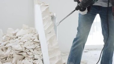 Kırmızı kareli kotlar ve koruyucu eldivenler giyen kimliği belirsiz erkek inşaat işçisi, döner çekiç matkabıyla beyaz duvarı yıkıyor, yerde enkaz oluşturuyor, manzarayı kapatıyor. Yenileme.