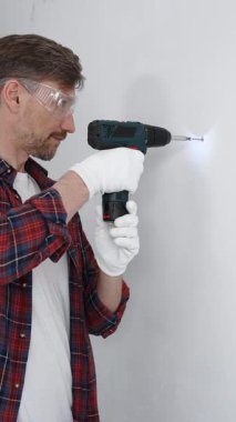 Kırmızı tişört ve beyaz eldiven giyen orta yaşlı erkek işçi, ev yenileme çalışmaları sırasında kablosuz matkapla duvarda delik açıyor. Dikey görünüm.