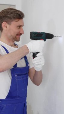 Mavi üniformalı profesyonel işçi, güvenlik eldivenleri, tadilat sırasında beyaz duvarda delik açmak için kablosuz matkap kullanıyor. Dikey portre görünümü.