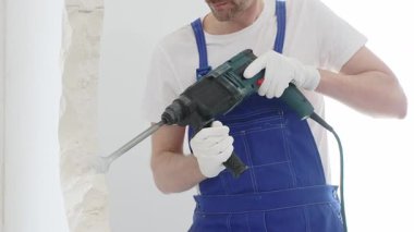 Mavi tulumlu ve koruyucu beyaz eldivenli tanımlanamayan erkek inşaat işçisi, beyaz duvarı döner çekiç matkapıyla yıkıyor, toz üretiyor ve yatay görüşü kapatıyor. Yenileme