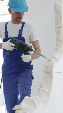 Mavi tulumlu ve koruyucu beyaz eldivenli bir inşaat işçisi, beyaz duvarı döner çekiç matkabıyla yıkıyor, toz üretiyor, yatay görüşü kapatıyor. Yenileme konsepti.