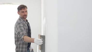 Koruyucu eldiven ve ekose gömlek giyen erkek inşaat işçisi, tadilat projesi sırasında profesyonel alçı levha bantlama bıçağı kullanarak duvarın yüzeyine sorunsuzca alçı yayıyor. Dikey görünüm.