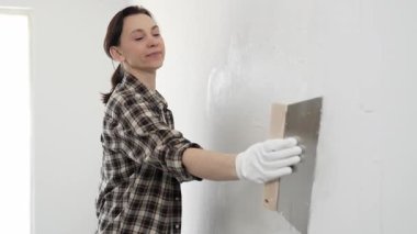 Kareli gömlek giyen kadın inşaat işçisi, beyaz duvar yüzeyini düzleştiriyor, profesyonel spatula tekniği ile macunu uyguluyor, iç dekorasyon konusunda yetenekli..