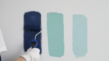 Eldiven giyen ressam beyaz bir duvara boya silindiri uyguluyor, mavi rengin çeşitli tonlarını test ederek iç tasarım için güzel bir eğim etkisi yaratıyor.