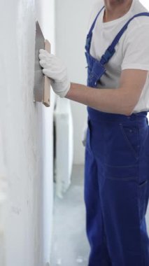 Koruyucu eldivenler ve mavi inşaat tulumları giyen erkek inşaat işçisi, tadilat projesi sırasında profesyonel alçı levha bantlı bıçak kullanarak duvarlara sorunsuzca alçı yayıyor. Portre