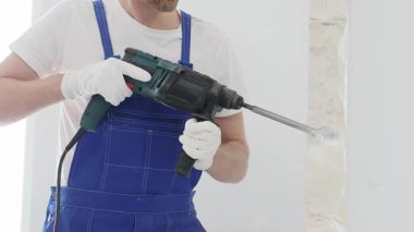 Mavi tulumlu ve koruyucu beyaz eldivenli tanımlanamayan erkek inşaat işçisi, beyaz duvarı döner çekiç matkapıyla yıkıyor, toz üretiyor ve yatay görüşü kapatıyor. Yenileme