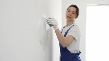 Profesyonel inşaat işçisi kadın, beyaz bir duvarı onarmak için macun bıçağı kullanıyor, bir kat macun sürüyor ve temiz bir iş için beyaz eldiven takıyor.