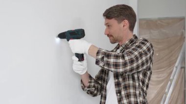 İşaretli gömlek ve beyaz eldiven giyen orta yaşlı bir adam, ev yenileme çalışmaları sırasında kablosuz matkapla duvarda delik açıyor. Dikey görünüm.
