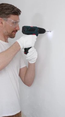 Beyaz tişört ve koruyucu eldiven giyen orta yaşlı erkek işçi, ev yenileme çalışmaları sırasında kablosuz matkapla duvarda delik açıyor. Dikey görünüm.
