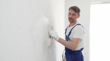 Koruyucu eldivenler ve mavi inşaat tulumları giyen erkek inşaat işçisi, tadilat projesi sırasında profesyonel alçı levha bantlı bıçak kullanarak duvarlara sorunsuzca alçı yayıyor. Portre
