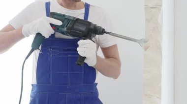 Mavi tulumlu ve koruyucu beyaz eldivenli tanımlanamayan erkek inşaat işçisi, beyaz duvarı döner çekiç matkapıyla yıkıyor, toz üretiyor ve yatay görüşü kapatıyor. Yenileme