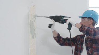 Kırmızı kareli gömlek, mavi başlık ve koruyucu eldiven giyen inşaat işçisi, döner çekiç matkabıyla beyaz duvarı yıkıyor, toz ve portre görüntüsü yaratıyor. Yenileme konsepti.