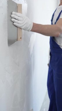 Koruyucu eldivenler ve mavi inşaat tulumları giyen erkek inşaat işçisi, tadilat projesi sırasında profesyonel alçı levha bantlı bıçak kullanarak duvarlara sorunsuzca alçı yayıyor. Portre