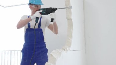 Mavi tulumlu ve koruyucu beyaz eldivenli bir inşaat işçisi, beyaz duvarı döner çekiç matkabıyla yıkıyor, toz üretiyor, yatay görüşü kapatıyor. Yenileme konsepti.