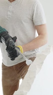 Beyaz tişörtlü ve koruyucu eldivenli, kimliği belirsiz bir inşaat işçisi, beyaz duvarı döner çekiç matkapıyla yıkıyor, yerde enkaz oluşturuyor, dikey yakın görüş açısında. Yenileme.