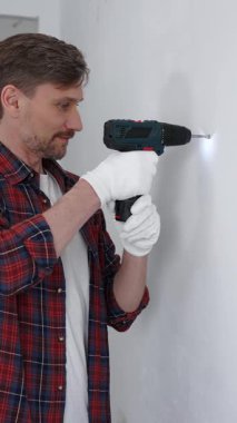 Kırmızı tişört ve beyaz eldiven giyen orta yaşlı erkek işçi, ev yenileme çalışmaları sırasında kablosuz matkapla duvarda delik açıyor. Dikey görünüm.