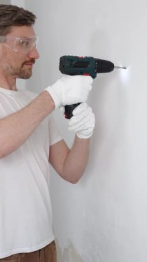 Beyaz tişört ve koruyucu eldiven giyen orta yaşlı erkek işçi, ev yenileme çalışmaları sırasında kablosuz matkapla duvarda delik açıyor. Dikey görünüm.