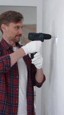 Kırmızı tişört ve beyaz eldiven giyen orta yaşlı erkek işçi, ev yenileme çalışmaları sırasında kablosuz matkapla duvarda delik açıyor. Dikey görünüm.