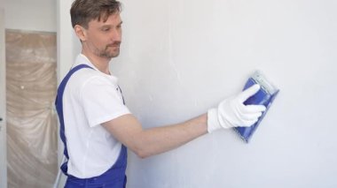 Koruyucu beyaz eldiven ve mavi inşaat tulumları giyen inşaat işçisi, iç dekorasyon projesi sırasında pürüzsüz bir yüzey hazırlamak üzere mavi el bloğuyla beyaz duvarı zımparalıyor..