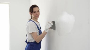 Profesyonel inşaat işçisi kadın, beyaz bir duvarı onarmak için macun bıçağı kullanıyor, bir kat macun sürüyor ve temiz bir iş için beyaz eldiven takıyor.
