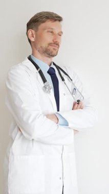 Kendine güvenen erkek doktor kolları çapraz duruyor, steteskop ve beyaz önlük giyiyor. Düşünceli bir şekilde, tıp alanında profesyonelliği ve uzmanlığı somutlaştırıyor. İlaç ve sağlık hizmetleri