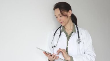 Yeşil bluz, steteskop üzerine beyaz önlük giyen doktor kadın dijital tablet üzerindeki hasta kayıtlarını inceliyor. Tıbbi çalışma alanında doğal ışıklandırmayla duruyor. Tıp konsepti.