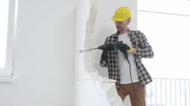 Bej kareli gömlek, sarı başlık ve koruyucu eldiven giyen inşaat işçisi, dönen çekiç matkabıyla beyaz duvarı yıkıyor ve orta uzunlukta toz üretiyor. Yenileme konsepti