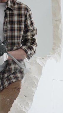 Tanınmayan bej kareli gömlek ve koruyucu eldivenler giyen erkek inşaat işçisi, döner çekiç matkabıyla beyaz duvarı yıkıyor, toz üretiyor, yakın görüş sağlıyor. Yenileme konsepti.