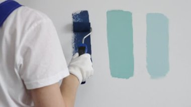 Eldiven giyen ressam beyaz bir duvara boya silindiri uyguluyor, mavi rengin çeşitli tonlarını test ederek iç tasarım için güzel bir eğim etkisi yaratıyor.