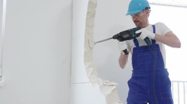 Mavi tulumlu ve koruyucu beyaz eldivenli bir inşaat işçisi, beyaz duvarı döner çekiç matkabıyla yıkıyor, toz üretiyor, yatay görüşü kapatıyor. Yenileme konsepti.