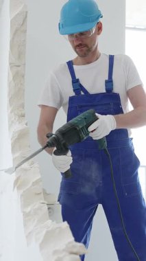 Mavi tulumlu ve koruyucu beyaz eldivenli bir inşaat işçisi, beyaz duvarı döner çekiç matkabıyla yıkıyor, toz üretiyor, yatay görüşü kapatıyor. Yenileme konsepti.