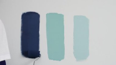 Eldiven giyen ressam beyaz bir duvara boya silindiri uyguluyor, mavi rengin çeşitli tonlarını test ederek iç tasarım için güzel bir eğim etkisi yaratıyor.