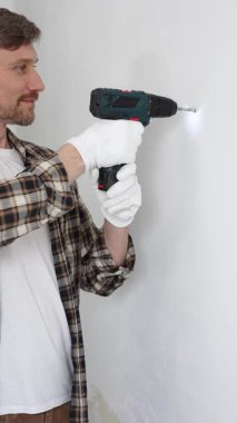 İşaretli gömlek ve beyaz eldiven giyen orta yaşlı bir adam, ev yenileme çalışmaları sırasında kablosuz matkapla duvarda delik açıyor. Dikey görünüm.