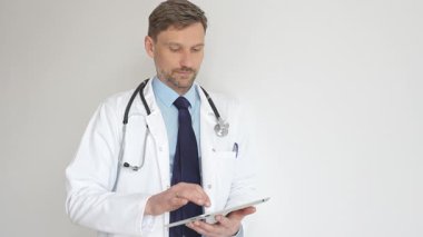 Beyaz önlüklü ve steteskoplu bir doktor dijital tablet kullanıyor. Hasta kayıtlarını güncellemeye odaklanmış, modern sağlık teknolojisi ve profesyonel uzmanlığın altını çizmiş. İlaç ve sağlık hizmetleri