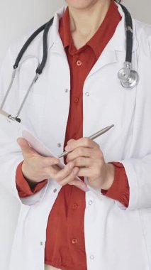 Tuğla kırmızı bluzun üzerine steteskop ve beyaz önlük giyen kimliği belirsiz kadın doktor tıp uzmanlığını ve hasta bakımını temsil eden bir panoya yazı yazıyor. Tıp konsepti.