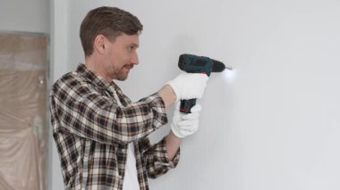 İşaretli gömlek ve beyaz eldiven giyen orta yaşlı bir adam, ev yenileme çalışmaları sırasında kablosuz matkapla duvarda delik açıyor. Dikey görünüm.