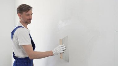 Koruyucu eldivenler ve mavi inşaat tulumları giyen erkek inşaat işçisi, tadilat projesi sırasında profesyonel alçı levha bantlı bıçak kullanarak duvarlara sorunsuzca alçı yayıyor. Portre