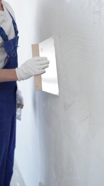 Koruyucu eldivenler ve mavi inşaat tulumları giyen erkek inşaat işçisi, tadilat projesi sırasında profesyonel alçı levha bantlı bıçak kullanarak duvarlara sorunsuzca alçı yayıyor. Portre