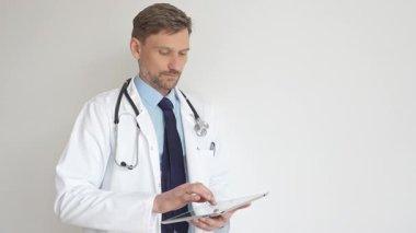 Beyaz önlüklü ve steteskoplu bir doktor dijital tablet kullanıyor. Hasta kayıtlarını güncellemeye odaklanmış, modern sağlık teknolojisi ve profesyonel uzmanlığın altını çizmiş. İlaç ve sağlık hizmetleri