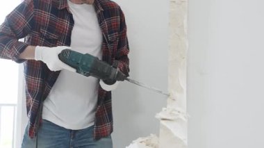 Kırmızı kareli kot pantolon ve koruyucu eldiven giyen kimliği belirsiz erkek inşaat işçisi, döner çekiç matkabıyla beyaz duvarı yıkıyor, toz üretiyor, yakın görüş sağlıyor. Yenileme konsepti.